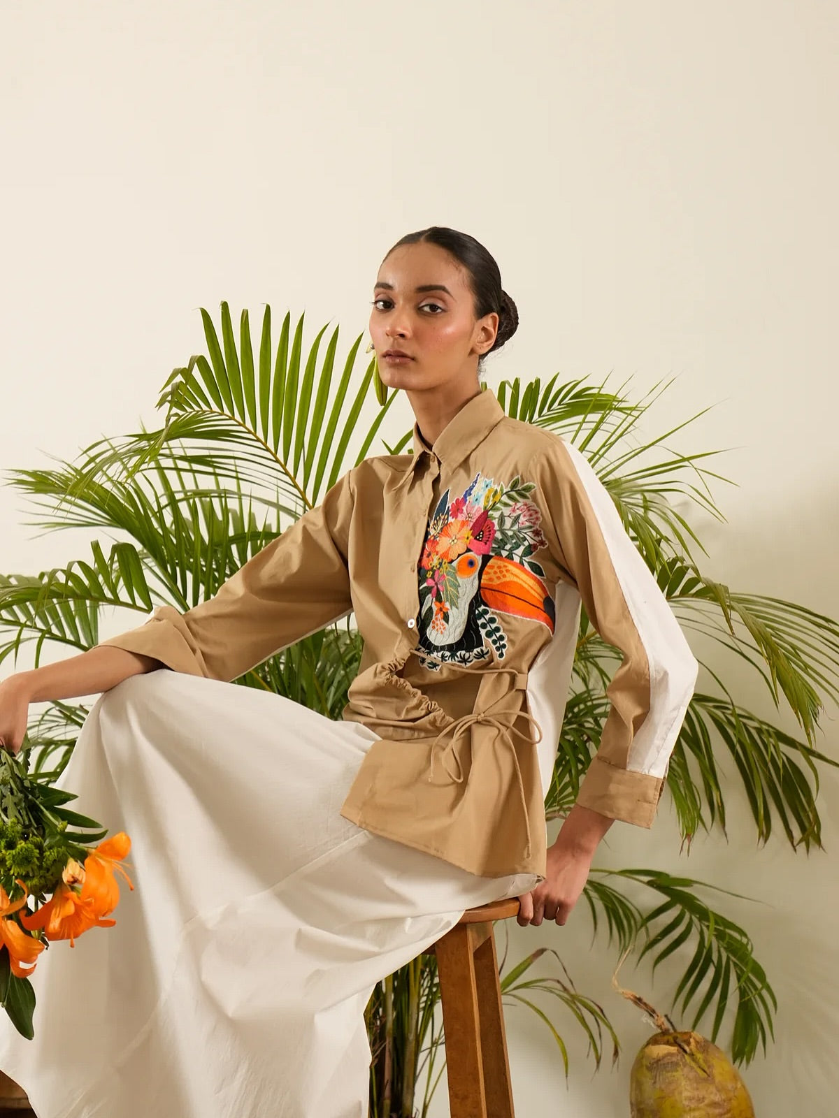 Woman on a stool in a printed shirt jacket beside palm fronds and lilies